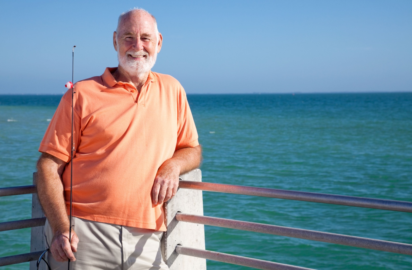 An older adult smiling while holding a fishing rod and leaning against the rail of a pier in front of a lake.