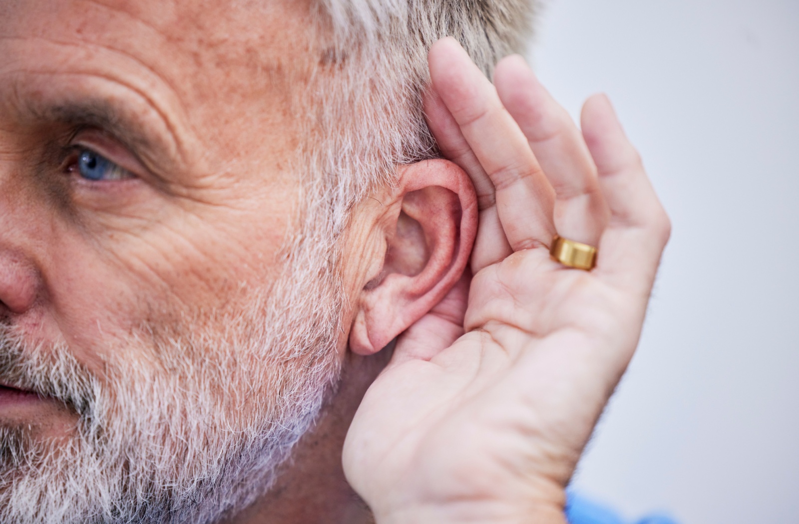 A senior wearing a golden wedding ring holds a hand up to their ear, trying to compensate for worsening hearing