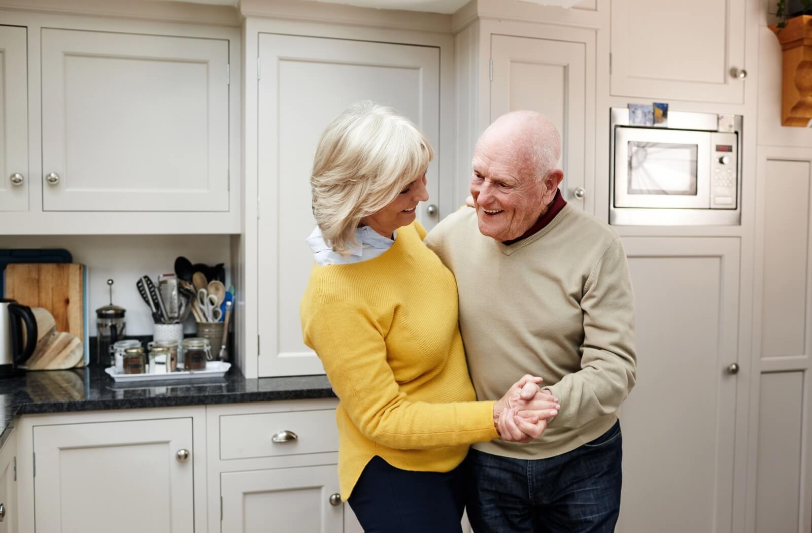 Senior couple dancing and smiling in a cozy kitchen with white cabinets and natural lighting
