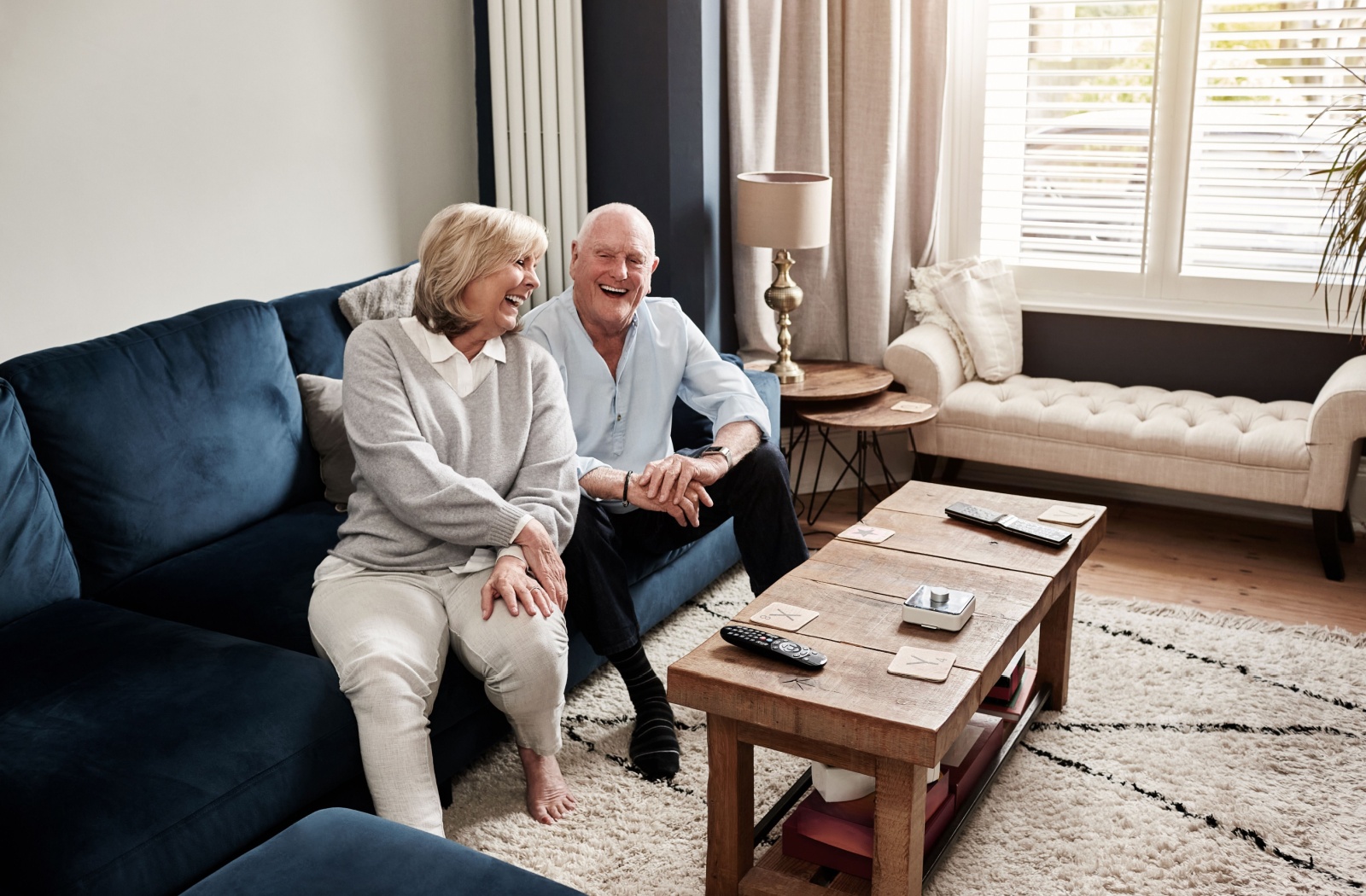 Senior couple laughing together on a navy blue couch in a bright living room with large windows and soft decor