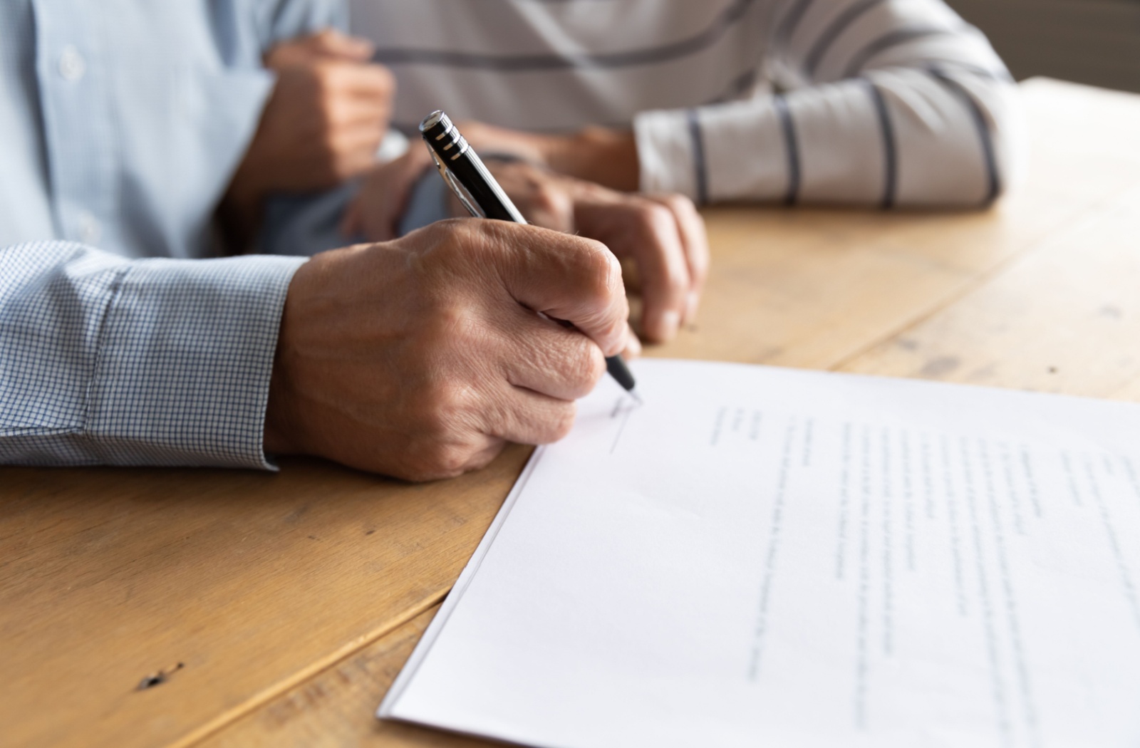 A close-up image of an older adult signing a legal document while their spouse holds their arm in support