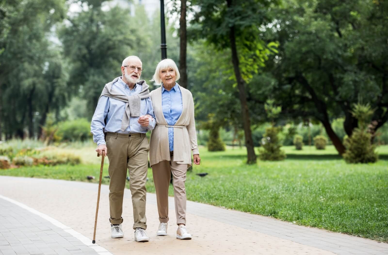 An older couple walks arm-in-arm along a pathway in a lush green park near their senior living community
