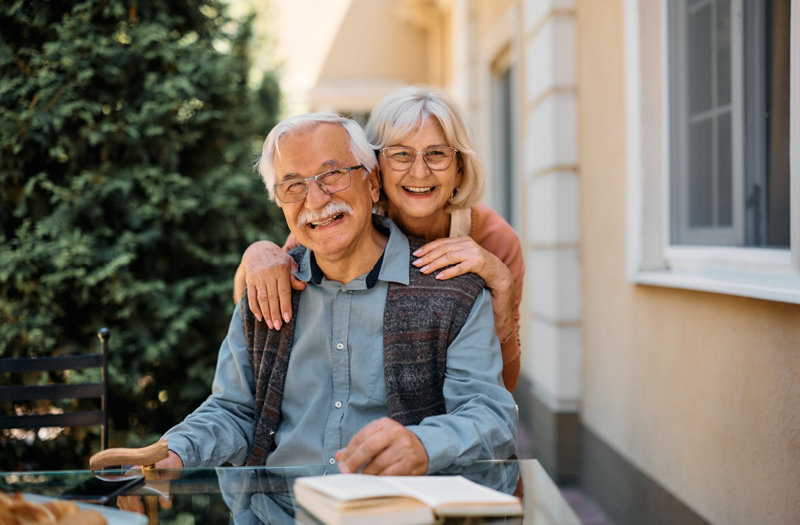 A smiling senior sits at a glass table outdoors while their spouse embraces them from behind