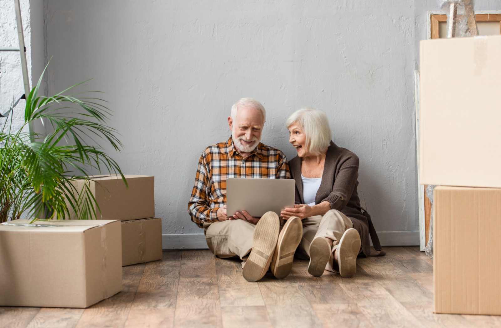 An older married couple sitting on the floor of their home surrounded by moving boxes, checking a moving list on a laptop.