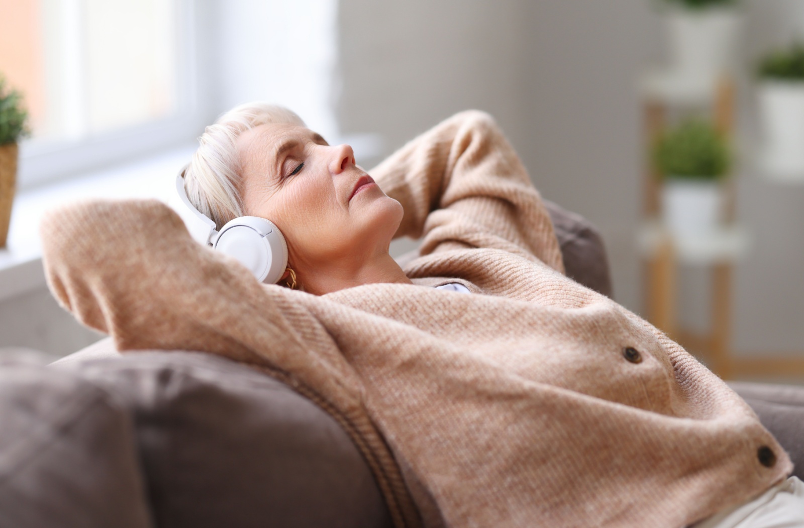 A senior wearing headphones relaxes with their eyes closed and listens to an audio book.