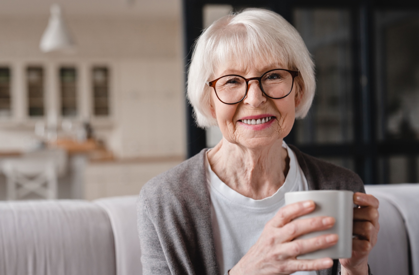 A senior woman with white hair holding a cup of tea, smiling and looking directly at the camera