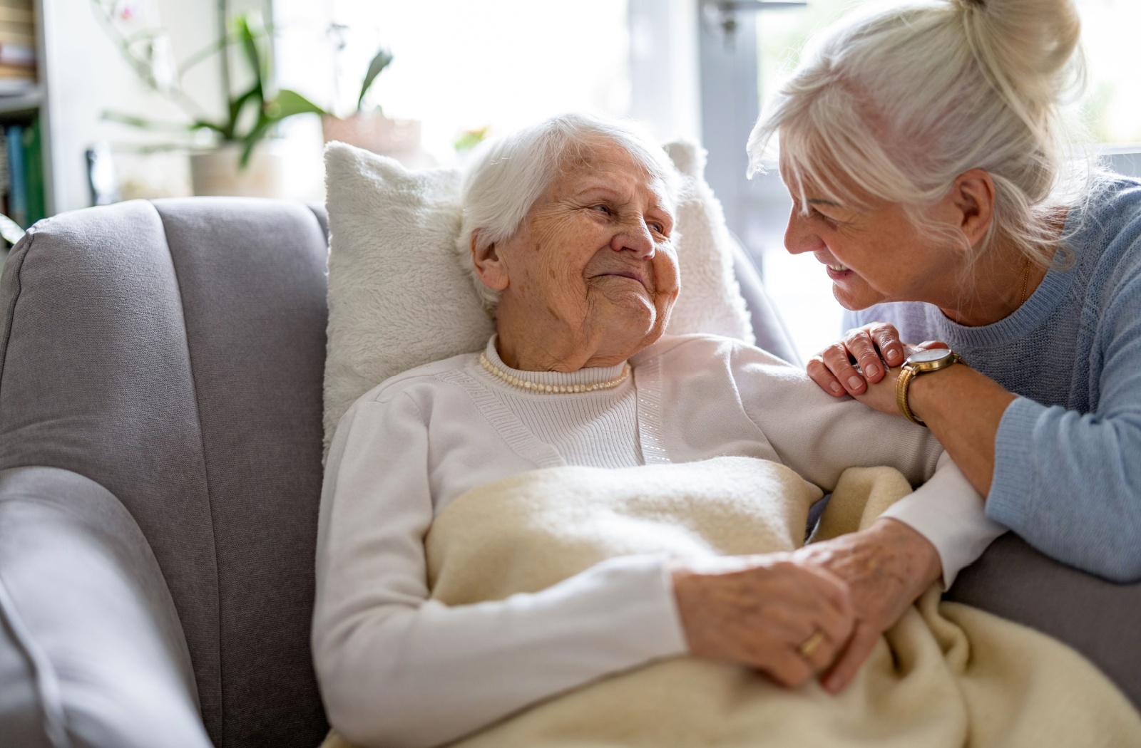 A smiling older adult sitting on a couch with a blanket looking at her smiling daughter beside her during a visit.