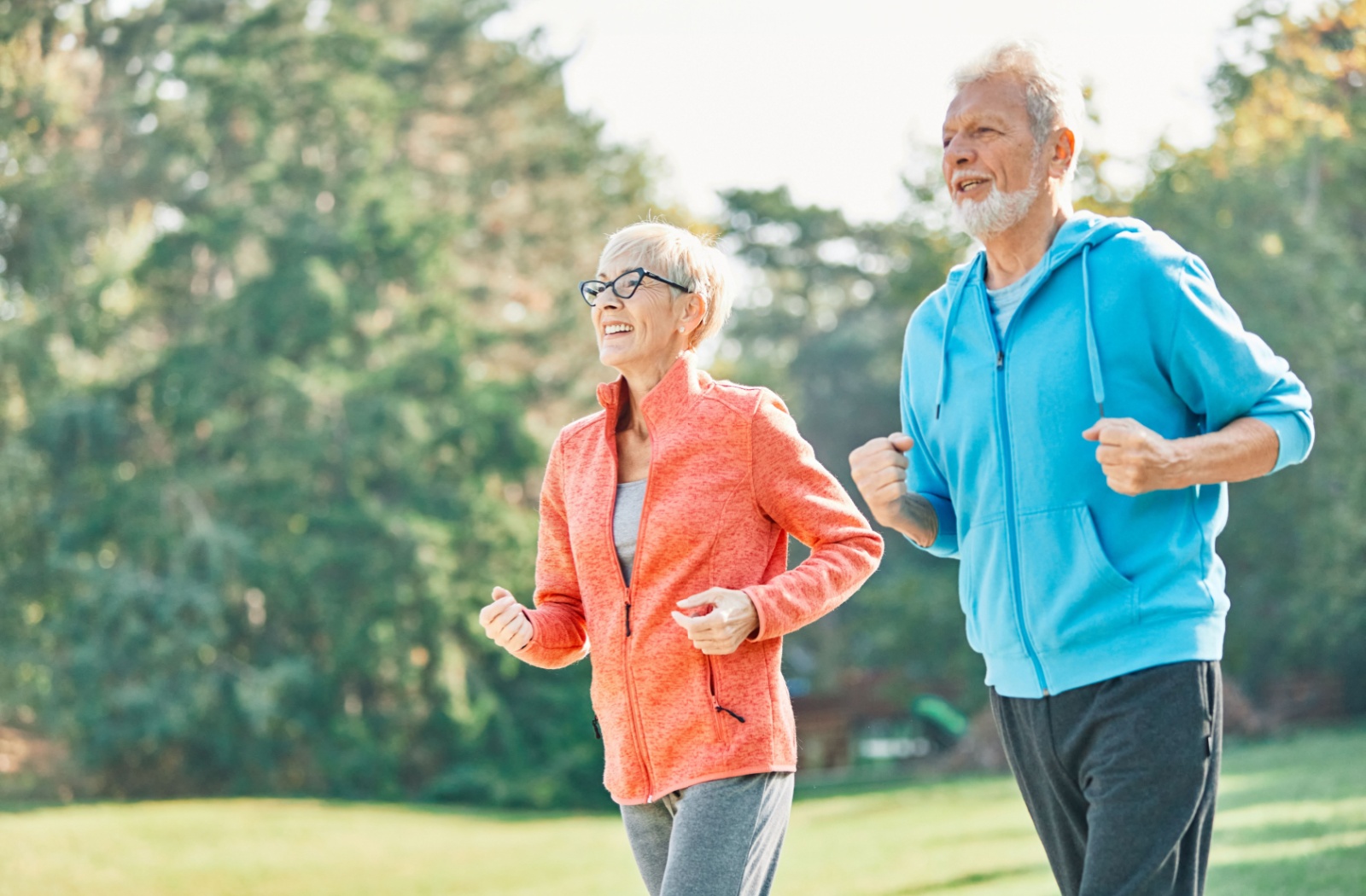 An older couple in exercise gear jogging outdoors in the park together to improve their cardiovascular health.