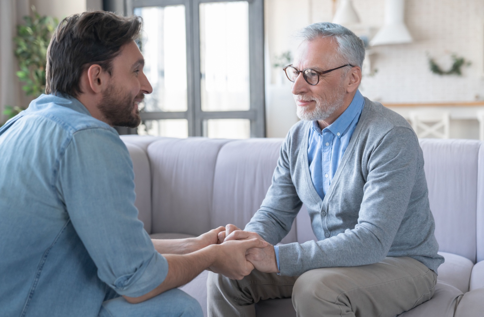 An adult son and his parent sitting across from each other holding hands during a conversation about assisted living.