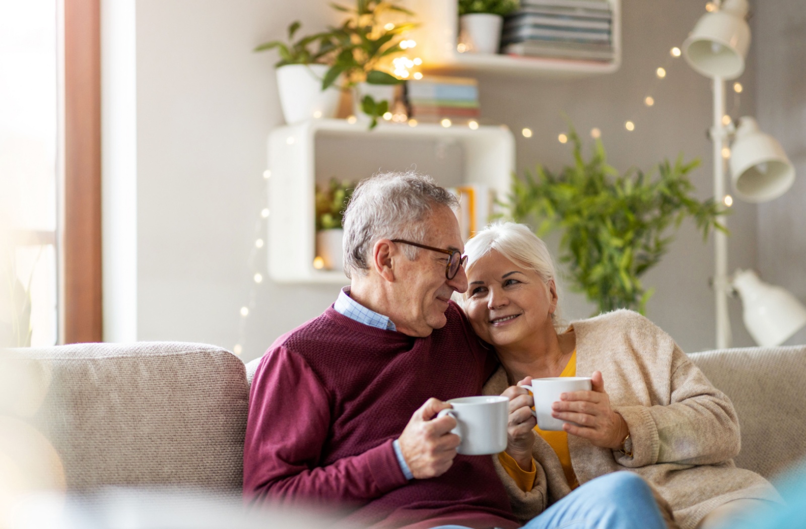 A smiling older couple holding mugs and sitting on the couch.