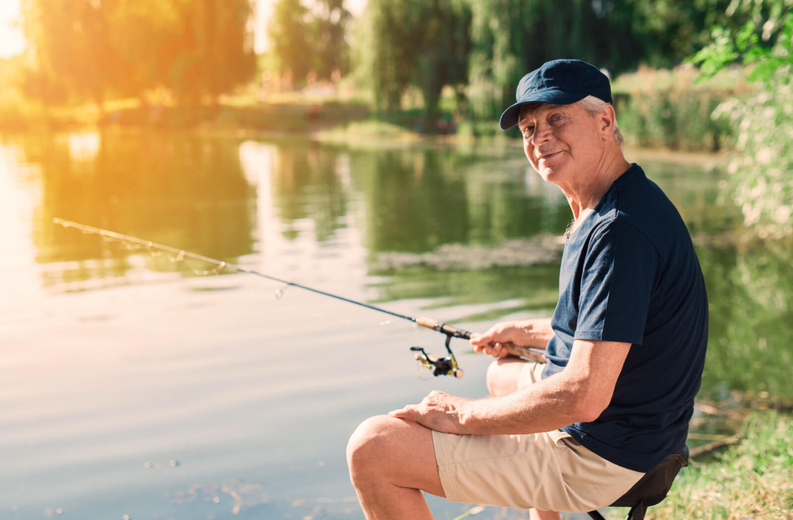 An older adult sitting on a small chair and fishing in a pond at sunrise.