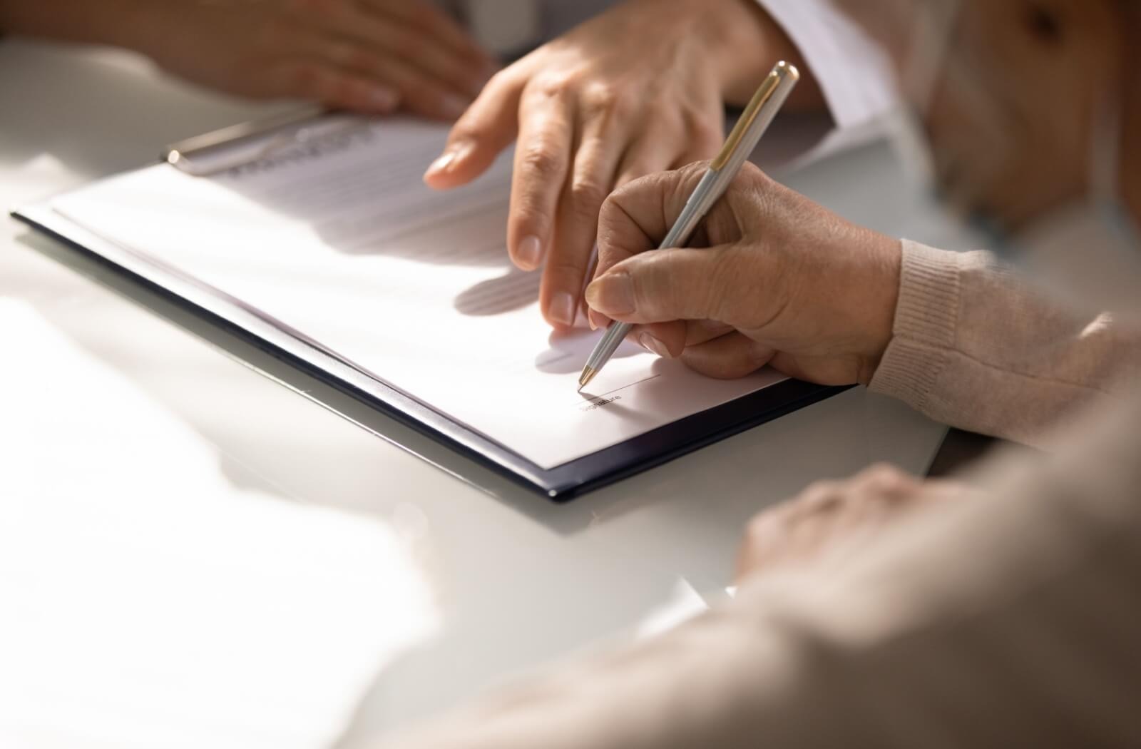 An older adult signs a legal document while an out-of-frame elder law attorney points to words on the page